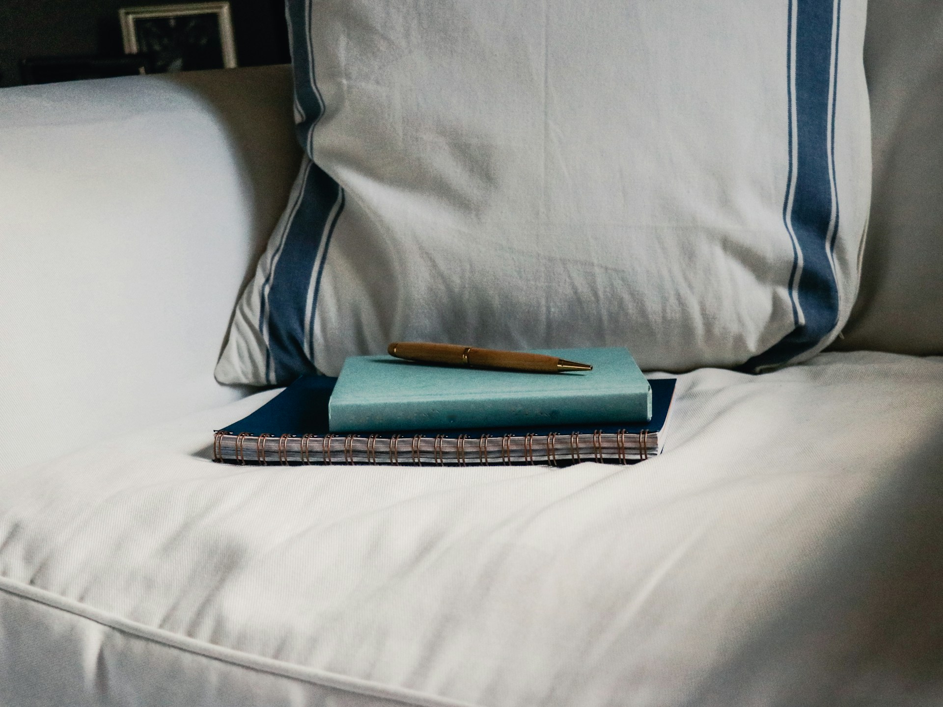 Two notebooks and a pen on a white armchair, in front of a white pillow with two blue stripes.